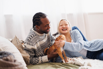 Joyful multiethnic young couple spending time with dog in bedroom