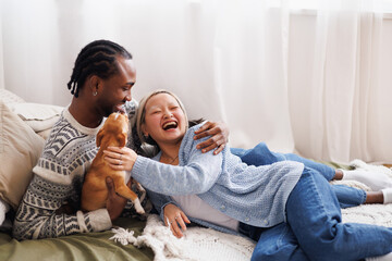 Smiling young interracial couple chilling with dog on bed at home