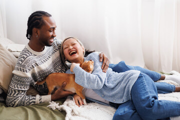 Cheerful young diverse couple relaxing with dog on bed at home