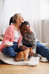 Joyful asian woman holding coffee and hugging african american boyfriend with pet at home
