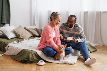 Positive relaxed multiethnic couple spending time with coffee and dog in bedroom