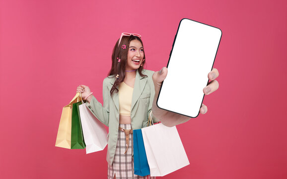 Trendy beautiful young Asian woman carrying colorful bags shopping online with mobile phone mockup of blank screen isolated on pink background.