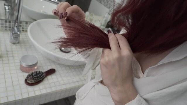 Closeup of a woman finds damaged hair, stands in front of a mirror, looks at dry hair tips, health care, medical procedures. Problem of split ends of hair