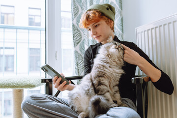 teenage girl sitting on chair, holding cat and looking at phone