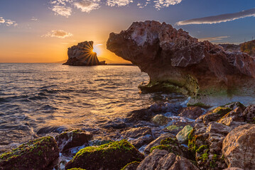 Rocks on the Peñon del Cuervo beach in Malaga, with the sun hidden behind the rock at sunset,...