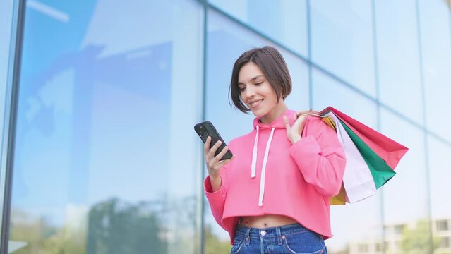 Positive caucasian female with telephone going home after shopping and looking at screen while sending message. Woman in cropped hoodie checking coupons and special offers in mobile app outdoor.