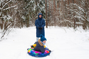 Fototapeta na wymiar Dad rides his son on an inflatable snow tube in the forest in winter. a man and his son are walking in a snowy park in winter.
