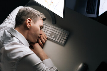 High-angle view of tired male trader asleep on computer desk late at night. Exhausted businessman sleeping with head on keyboard while working overnight on computer. Concept of overwork and deadline