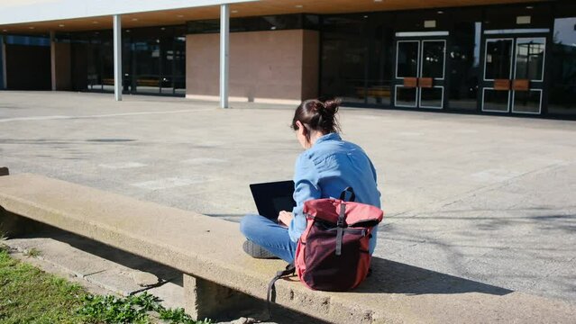 A young woman, university student, sits on campus, studying with her laptop, absorbed in learning and research, enjoying the tranquility outdoors.