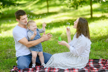 Obraz premium Happy family enjoying a picnic in the park in spring, blowing bubbles and smiling while sitting on a checkered blanket spread on the grass. Concept of a happy, playful, harmonious family relationship
