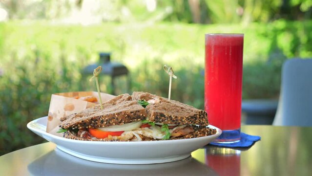 Outdoor brunch scene, fresh club sandwich on plate with side snacks and tall glass of red juice, served on table with natural backdrop. Casual dining and healthy eating.