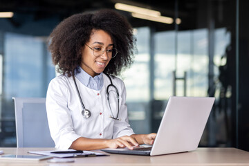Confident african american female doctor in medical uniform working by laptop in workspace. Smiling therapist with stethoscope prescribing medication after web consultation of client in clinic. - obrazy, fototapety, plakaty
