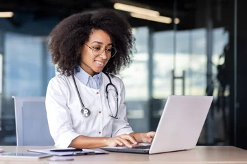 Poster Confident african american female doctor in medical uniform working by laptop in workspace. Smiling therapist with stethoscope prescribing medication after web consultation of client in clinic. © Liubomir