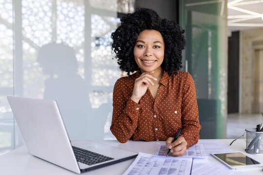 Latino woman in polka dot blouse filling out tables by hand while working at technically equipped desk. Motivated office administrator placing order to providing company with necessary supplies.