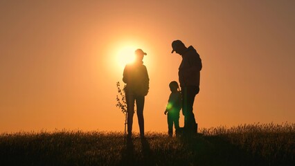 Mom dad child team planting tree in sun spring time. Family with tree at sunset Silhouette. Family with shovel, watering can plants young trees sprout in soil. Gardener dad mom child planting tree