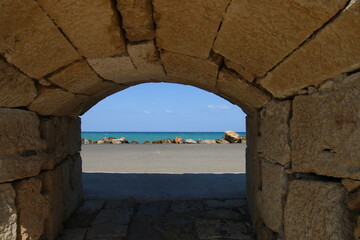 Arco de piedras a través del cual se ve en el fondo una carretera asfaltada, piedras y el mar con el cielo.