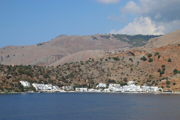 Pueblo costero de casas blancas junto a un mar azul y con una montaña marrón detrás, con algo de vegetación verde