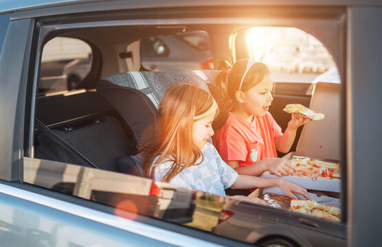 Two little sisters are happy to eat just cooked Italian pizza sitting in child car seats on the car back seat. Happy childhood, fast food eating, or auto journey lunch break concept image.