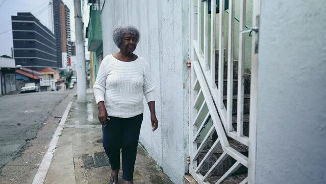 One elderly black lady opening residence front door, arriving home from urban sidewalk street. 80s African American woman returns to South American house after daily routine activity