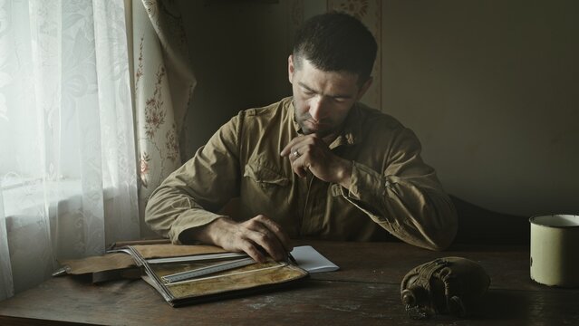 A Soviet Army soldier sits at a table in an abandoned farmhouse and looks at a map of the area. Reconstruction of the events of World War II intelligence soldier.
