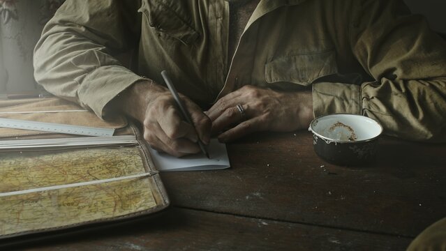 Soviet Army soldier analyzes map and draws up action plan sitting in abandoned house at table near window.