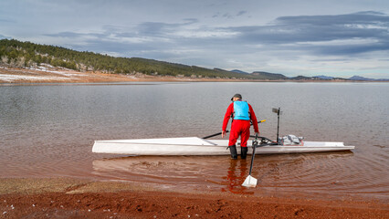 Senior male rower is launching a rowing shell on a shore of Carter Lake in northern Colorado in winter scenery.