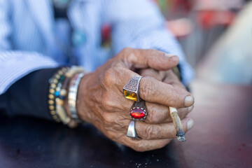 hands of an old man in a blue shirt with cheap jewelry, plastic stones and a cigarette on the...