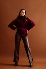 Fashionable confident woman wearing burgundy color knitted sweater, faux leather pants, pointed toe boots, posing on brown background. Full-length studio fashion portrait