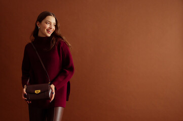 Fashionable happy smiling woman wearing burgundy color knitted sweater, carrying trendy leather...