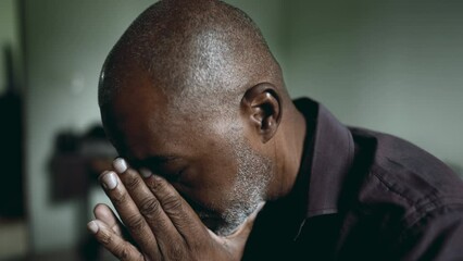 One depressed senior man covers face in shame and regret in moody gloomy bedroom. African American person struggles with poverty and mental illness, close-up face feeling hopeless