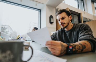 A pensive tattooed young man with a beard carefully examines paperwork in a bright, contemporary office setting, suggesting focus and professionalism.