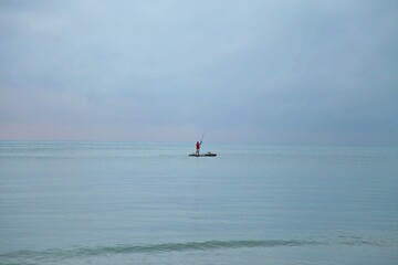 Solitude an fisherman alone over a raft in  wide blue sea.  Emptyness in early day, foggy sky mixing color with sea on horizon line