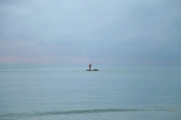 Fototapeta premium Solitude an fisherman alone over a raft in wide blue sea. Emptyness in early day, foggy sky mixing color with sea on horizon line