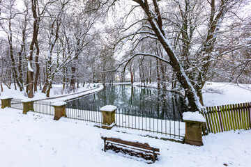 Winter snowy landscape. A brick bridge leading over a calm river, which is lined with tall snow-covered trees that are reflected on the surface of the river. A small bench in the foreground