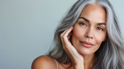 close-up portrait of an elegant senior woman with gray hair, gently touching her face and smiling subtly, set against a neutral background, suggesting a skincare or beauty theme.