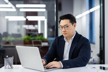 Serious concentrated asian businessman inside office at workplace, experienced mature boss in business suit typing on keyboard, thinking man.