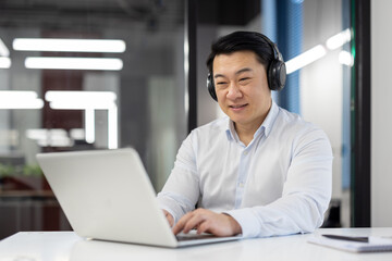 Successful asian programmer with laptop inside office, mature experienced man in headphones...