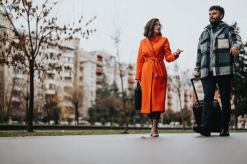 A fashionable couple strolls along an urban street carrying luggage, implying a casual city break...