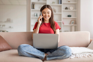 Young asian woman sitting on sofa with her laptop and talking phone