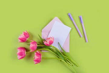 Bouquet of vibrant pink tulips beside empty greeting card and envelope on pastel green background. Top view, flat lay