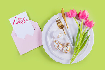 Pink tulips and table place setting with greeting or invitation card with inscription Happy Easter and envelope on pastel green background. Happy Easter card. Top view, flat lay