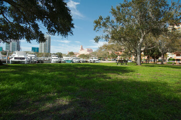 Low angle horizontal view over green grass at Vinoy Park in St. Petersburg, FL. Trees in foreground and sides with boats in water and buildings with blue and white sky later afternoon sun.