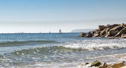 Fototapeta na wymiar View of the Mediterranean Sea, waves. In the background are ships and sailboats. sunny day. large stones on the shore