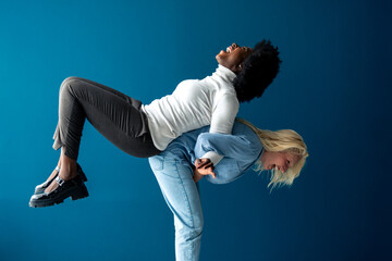 Interracial best friends laughing and having a good time together in a studio. Happy young women enjoying themselves while being playful against a blue background.