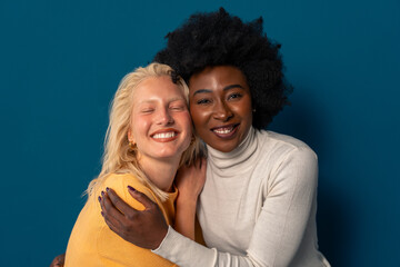 Young women of different ethnicity smiling happily while standing together against a studio background.
