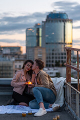 Surprise date on rooftop with urban cityscape and skyscrapers on background. Happy young loving couple drinking wine having romantic candlelit dinner celebrating anniversary or Valentines Day