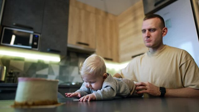 Blond baby boy lies on the table trying to get to the cake in front of him. Smiling dad sitting beside holds a kid.
