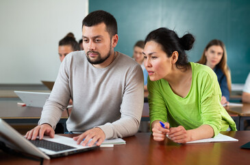 Asian woman and caucasian man university students sitting at table during lesson and using laptop.