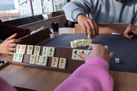 Group of friends sitting at wooden table and playing on Turkish board game Okey (Rummikub). Group game activity. People having fun while playing board game.