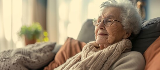 The elderly woman is sitting comfortably on a wooden couch in a cozy room, wearing glasses, a scarf, and a happy smile on her face.